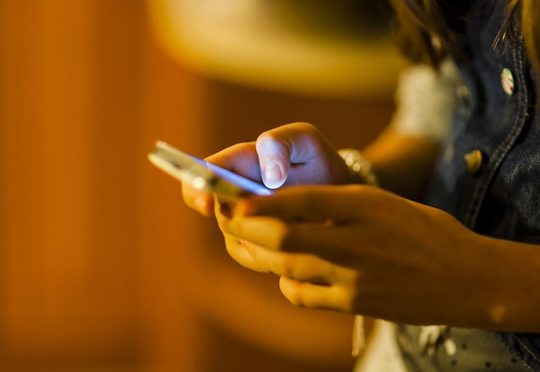 Woman in denim jacket holding a smart phone in her hands casting a cool light against a background lit with warm light