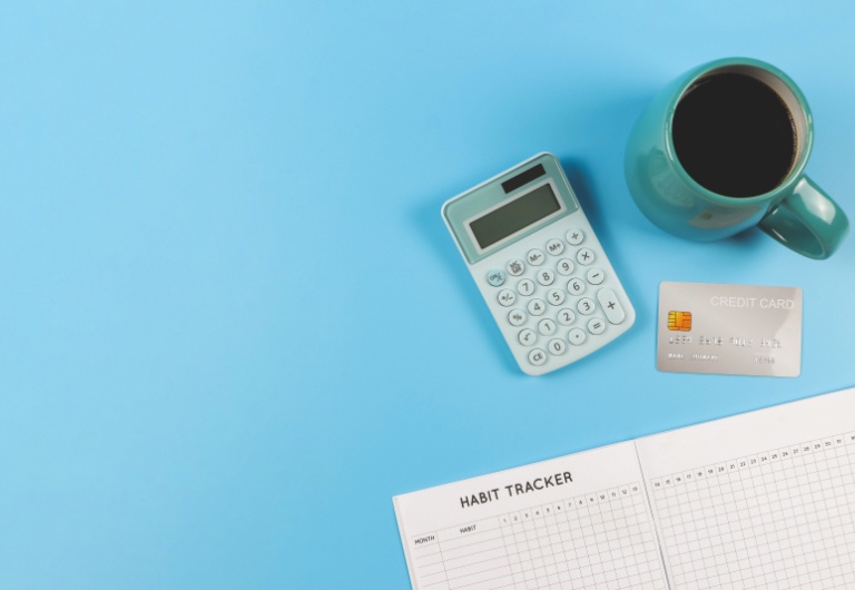 Top view or flat lay of habit tracker book, blue cup of black coffee, blue calculator and credit card on blue background with copy space