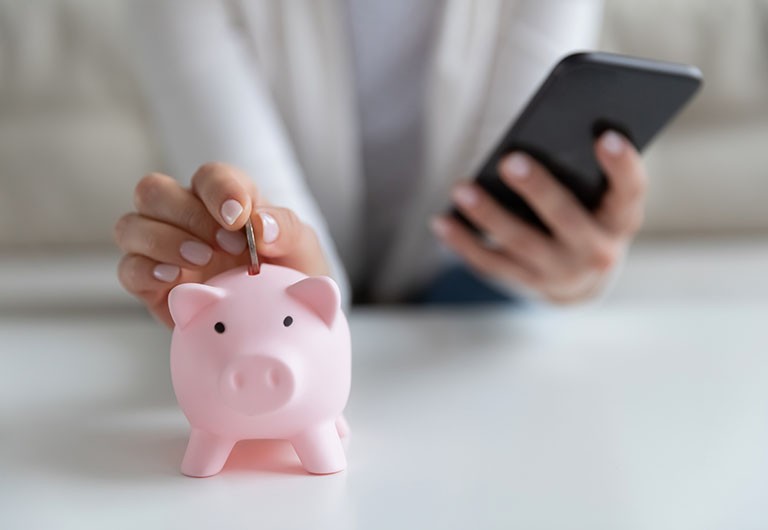 Hand placing coin into piggy bank while other hand holds smartphone